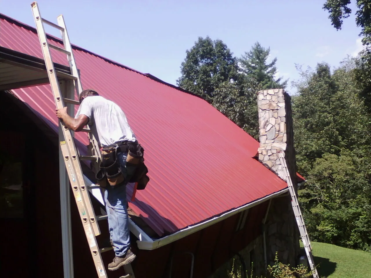 Roofer on a ladder beside a red metal roof and stone chimney — Soffit & Fascia Repair by Roofing Brothers in Moundridge
